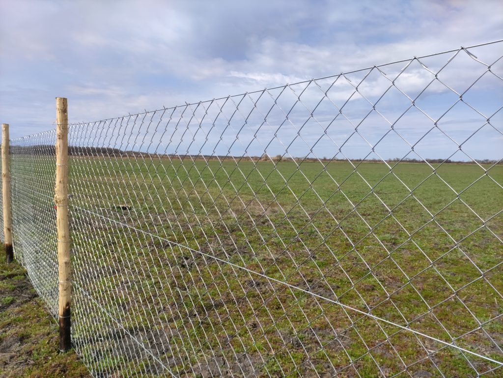 Drótkerítés 4 sor vezérdróttal, akác oszlopokkal, háttérben egy szántóföld.
