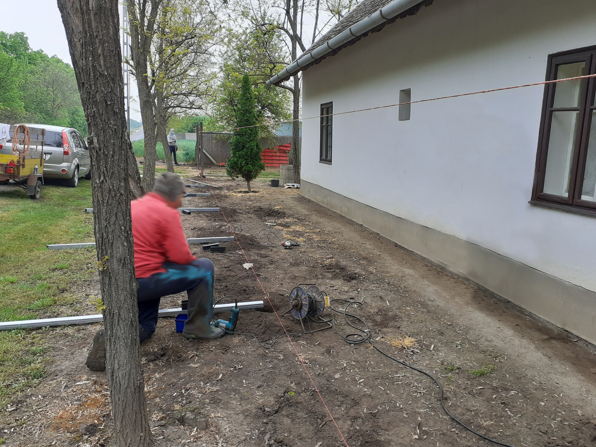 Régi ház frontján kerítésépítés zajlik, az oszlopok a fúratok elé fektetve.